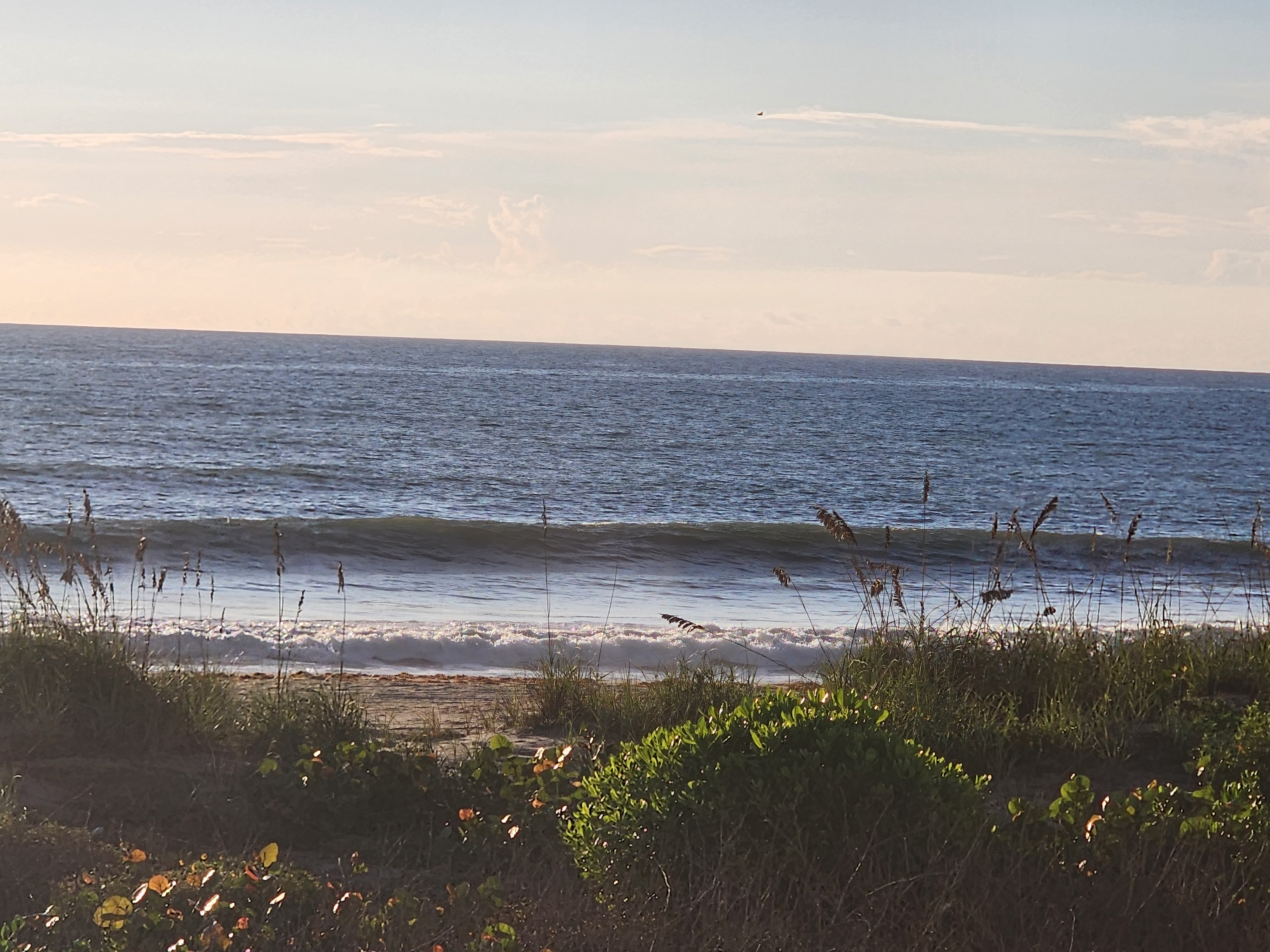 A wider dark line of swell pushing in toward the sand under morning haze, South Cocoa Beach