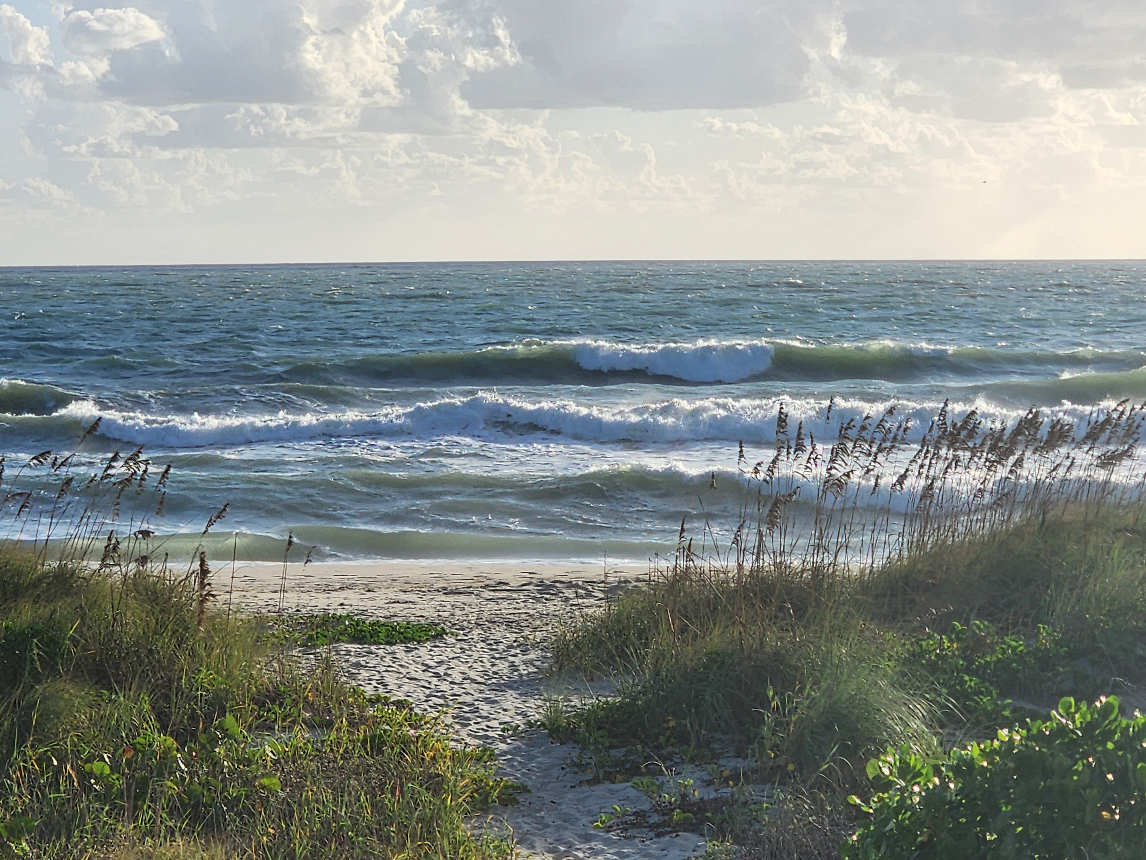 Multiple rows of whitewater and breaking waves rolling toward the shore at the end of the dune path, sea oats bending in the wind